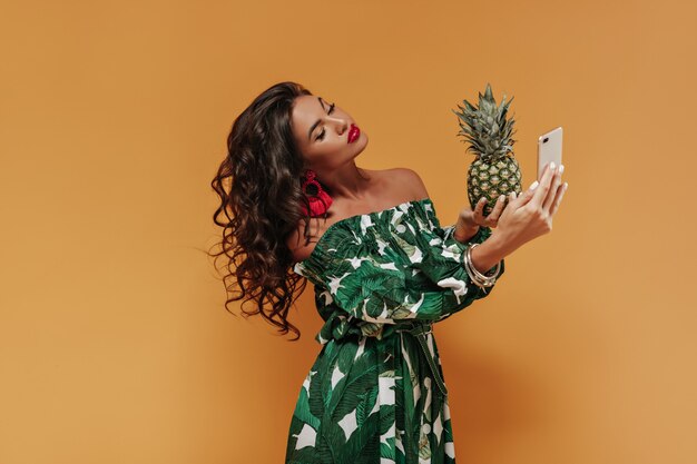 Curly modern girl with dark long hair and big lips in red earrings and printed summer dress making selfie and holding pineapple