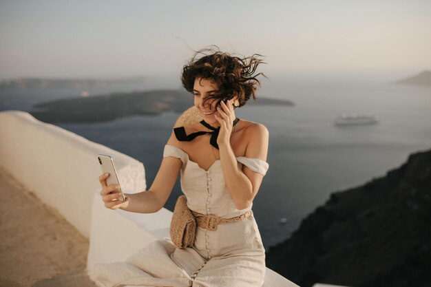 Curly shorthaired woman in beige dress smiles widely and takes selfie on sea background Charming brunette lady in stylish outfit sits on fence and holds phone