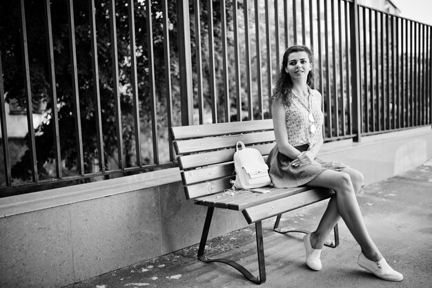 Curly stylish girl wear on blue jeans skirt blouse sitting on bench Portrait on streets of city