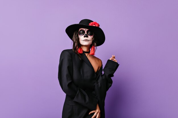 Curly stylish girl with red earrings and rose on black wide-brimmed hat posing pathetically in outfit for Halloween.