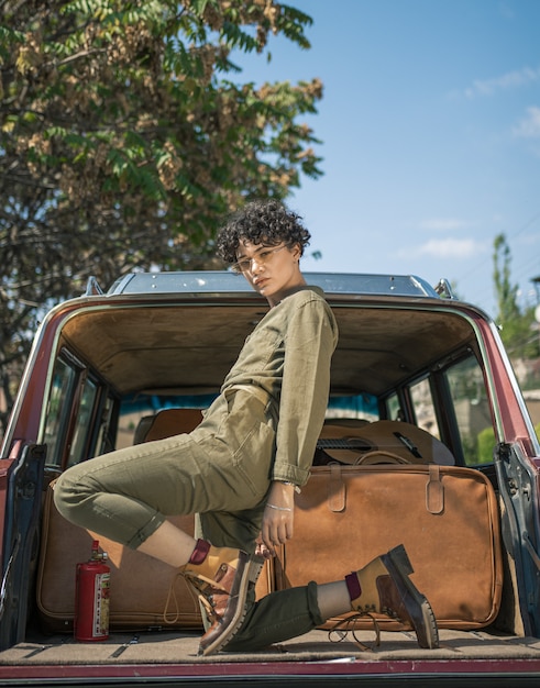 Curly stylish model posing for a photshot on the back of a car on a bright sunny day