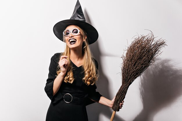 Curly witch in glasses expressing happiness in halloween. Indoor photo of laughing pretty girl in wizard costume holding broom.