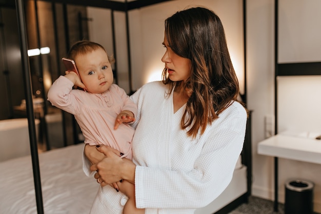 Curly woman in bathrobe looks puzzled at her little daughter, putting phone to her ear. Portrait of mom and baby in bright bedroom.