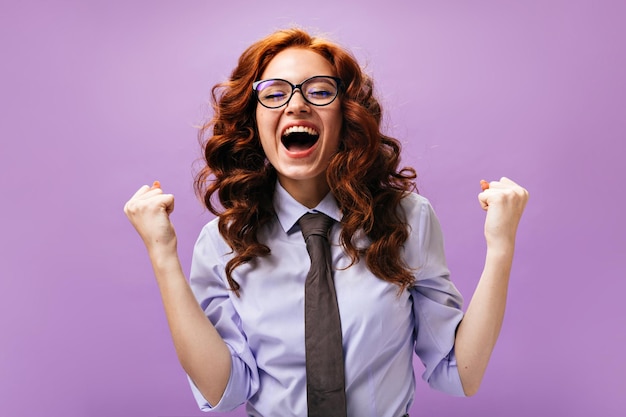 Кудрявая женщина в синей рубашке и очках счастливо позирует на фиолетовом фоне Веселая деловая девушка с рыжими волосами радуется