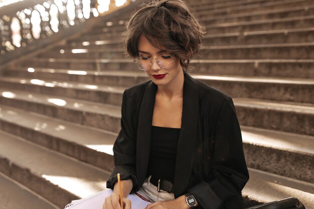 Curly woman in eyeglasses and black jacket writting outdoors. Beautiful woman with red lipstick and brunette hair sitting on stairs.