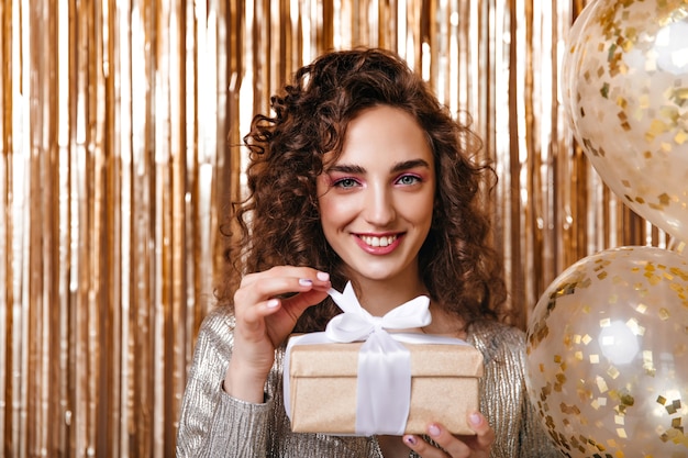 Curly woman in good humor holding gift box on golden background