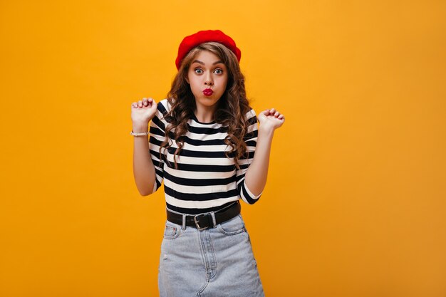 Curly woman in good mood makes funny face. Stylish girl in red beret and fashionable clothes has fun on isolated background.