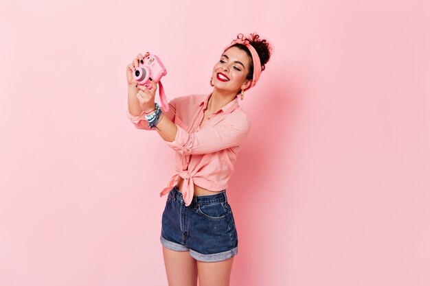 Curly woman in headband, pink blouse and denim shorts makes photo on mini camera on pink space.