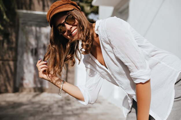 Curly woman is laughing. Friendly girl in white shirt and cap looks at camera on space of cozy houses.