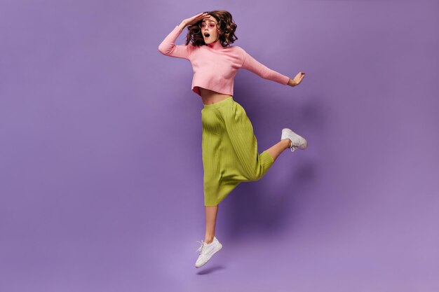 Curly woman jumping on purple background Young cheerful girl in green skirt and pink sweater looks away on isolated