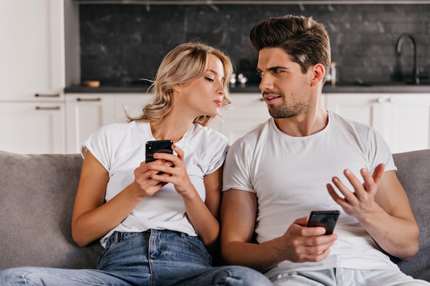 Curly woman looking at husband's phone. Appealing caucasian girl sitting on sofa with smartphone.