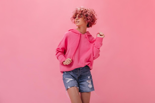 Curly woman in pink hoodie and denim shorts dance on isolated background Happy cool girl in sunglasses moves and smiles on pink backdrop