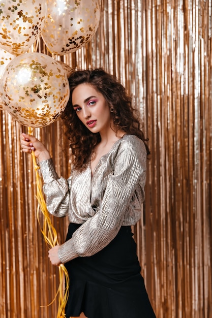 Curly woman in shiny top holding balloons on gold background