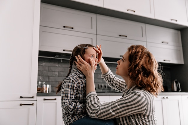 Curly woman in striped shirt carefully looks at her daughter and gently touches her face.