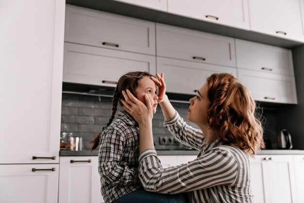 Curly woman in striped shirt carefully looks at her daughter and gently touches her face.