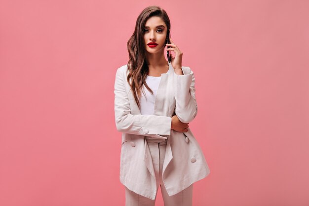 Curly woman in white suit talks on phone. Stylish lady with red lips in light fashionable outfit on pink isolated background.