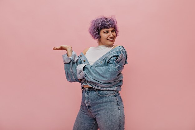 Curly woman with lilac hair in stylish denim suit posing on pink. Astonished woman looks away.