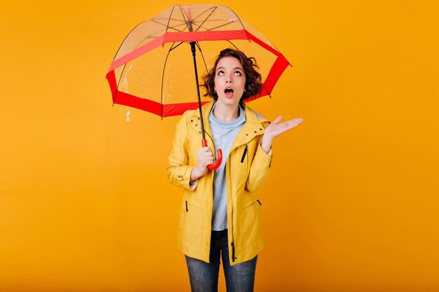 Curly woman in yellow coat expressing amazement standing under parasol.  portrait of emotional girl with umbrella, looking up with mouth open.