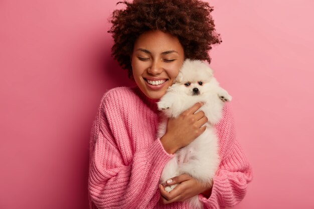 curly young woman embraces white spitz with love, being very happy to get present she dreamt about, wears oversized jumper, models against pink background.