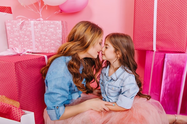 Curly young woman in retro outfit holding daughter's hands sitting on the floor next to present boxes and helium balloons. Portrait of birthday girl and her mom cute posing at the party with gifts