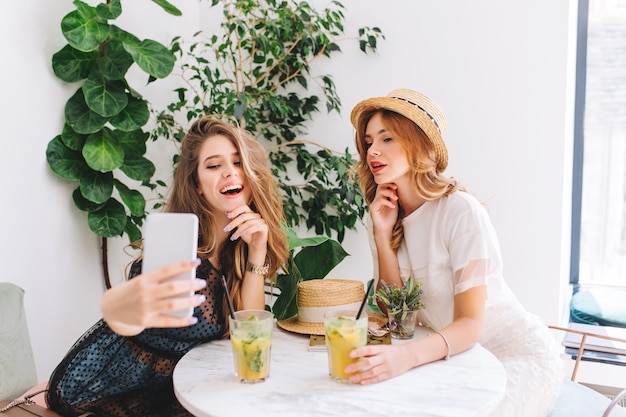 Curly young woman in straw hat holding glass of icy cocktail while her long-haired friend making selfie