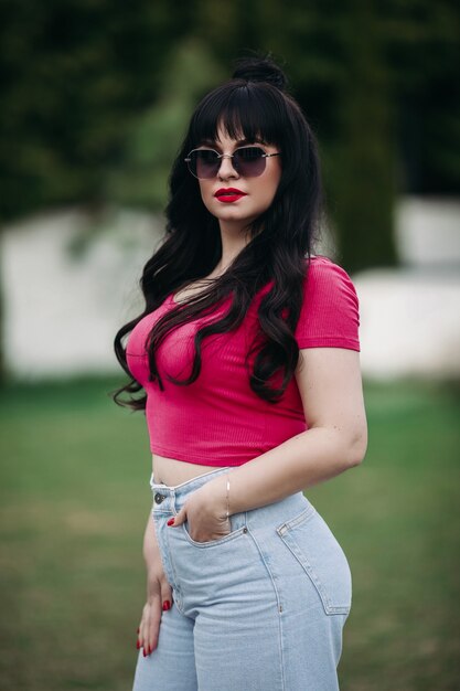 Curvy beautiful brunette woman wearing sunglasses and bright lipstick while standing outside on green lawn