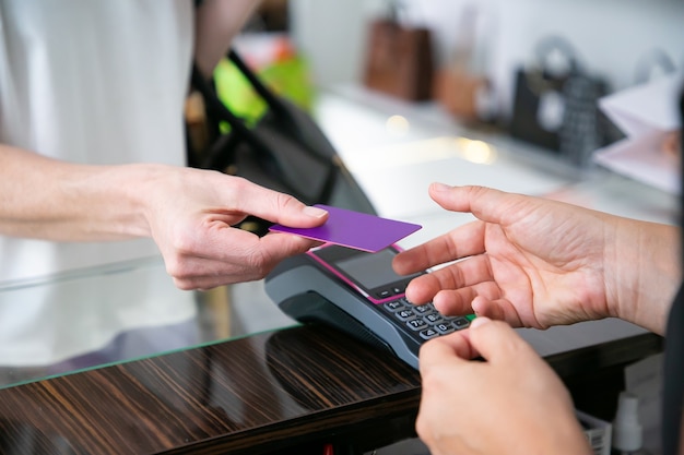 Customer giving credit card to cashier over desk with pos terminal for payment. Cropped shot, closeup of hands. Shopping concept