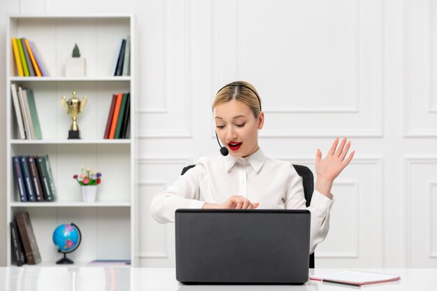 Customer service cute blonde girl in office shirt with headset and computer looking at clocks