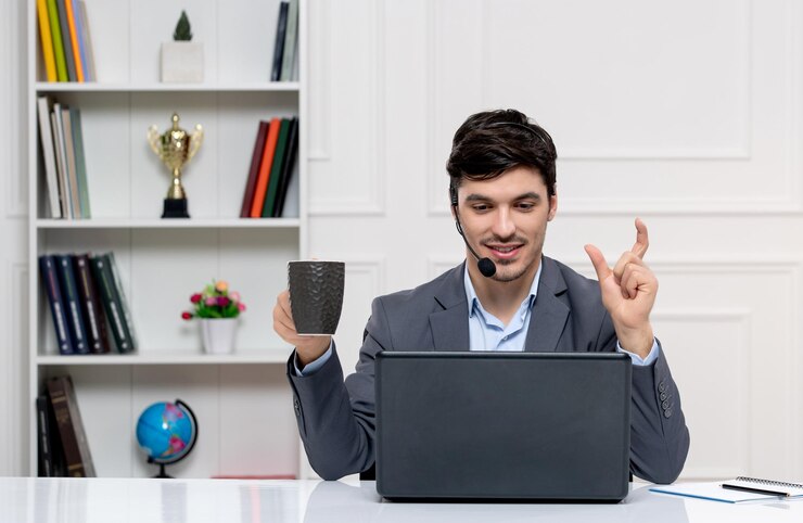 मुफ़्त फोटो customer service cute guy in grey suit with computer and headset holding a cup and smiling