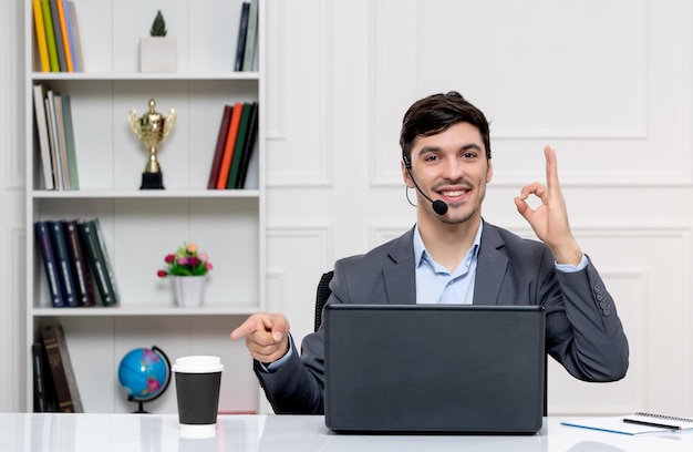 Foto gratuita servizio clienti ragazzo carino in abito grigio con computer e auricolare che mostra segno giusto e sorridente