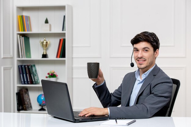 Customer service cute guy in grey suit with computer and headset smiling with coffee cup