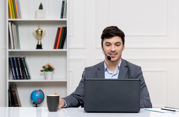 Customer service cute guy in grey suit with computer and headset talking on videocall