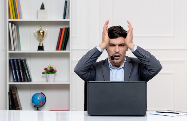 Customer service cute guy in grey suit with computer and headset very annoyed touching temples