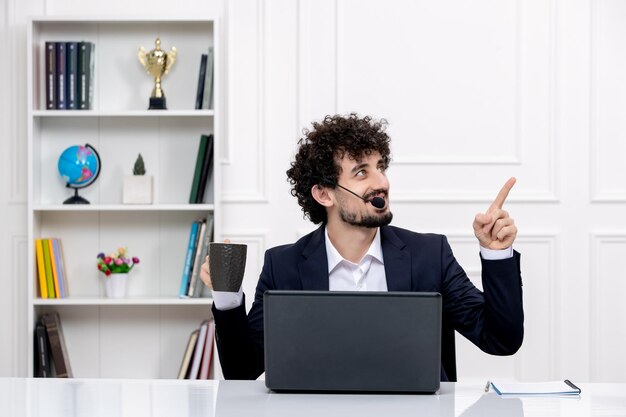 Customer service handsome curly man in office suit computer and headset looking up with coffee