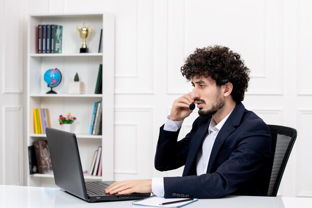 Customer service handsome curly man in office suit with computer and headset from the side