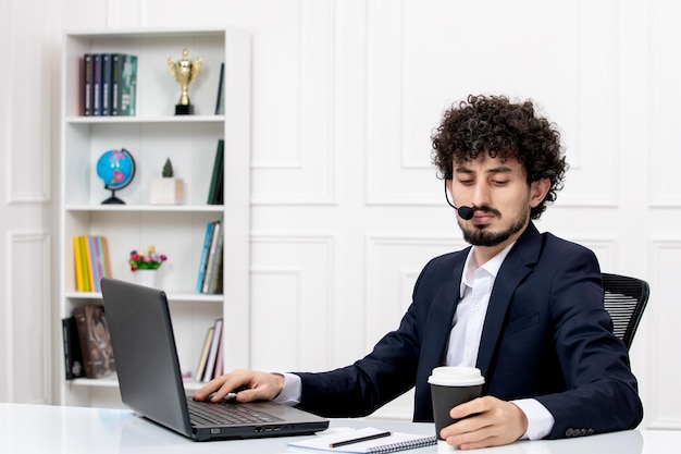 Customer service handsome curly man in office suit with computer and headset holding coffee
