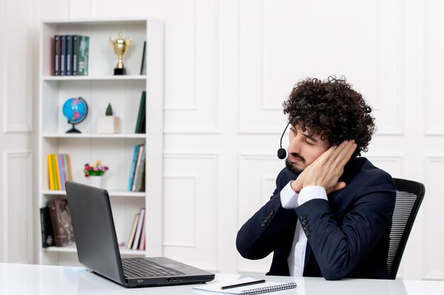 Customer service handsome curly man in office suit with computer and headset making sleep gesture