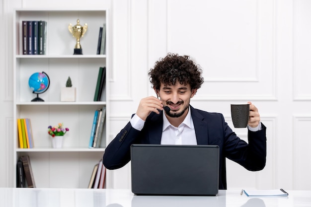 Servizio clienti bell'uomo riccio in tuta da ufficio con computer e auricolare sorridente con caffè