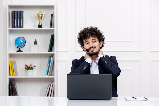 Customer service handsome curly man in office suit with computer and headset touching neck