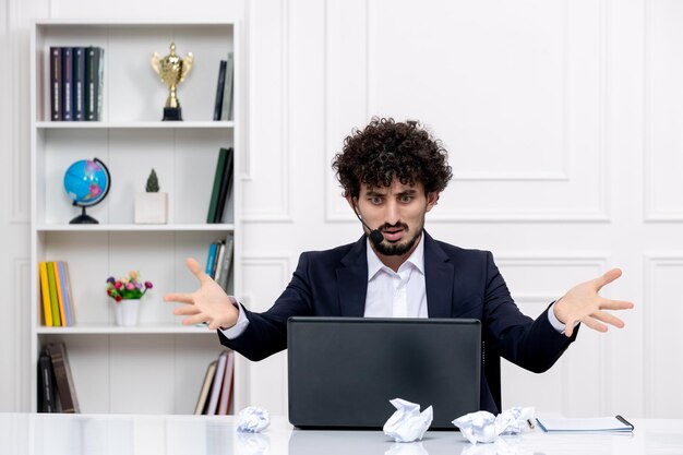 Customer service handsome curly man in office suit with computer and headset waving hands annoyed