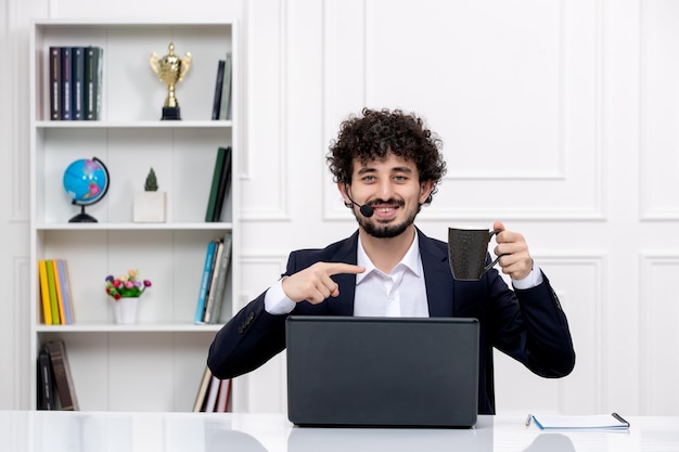 コンピューターとコーヒーとヘッドセットの幸せなオフィススーツのカスタマーサービスハンサムな巻き毛の男