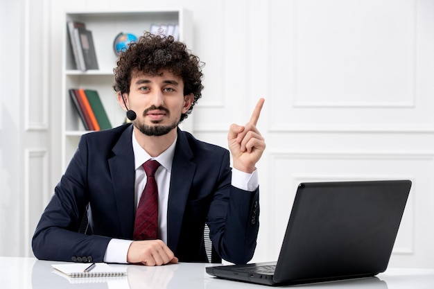 Customer service handsome young guy in office suit with laptop and headset pointing up