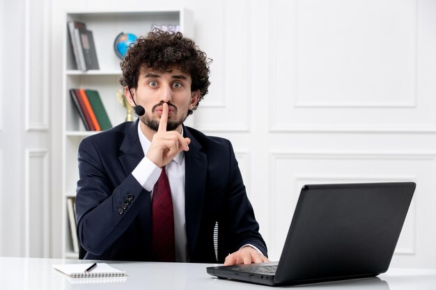 Customer service handsome young guy in office suit with laptop and headset showing silence sign