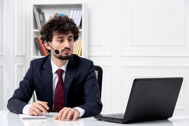 Customer service handsome young guy in office suit with laptop and headset taking down notes