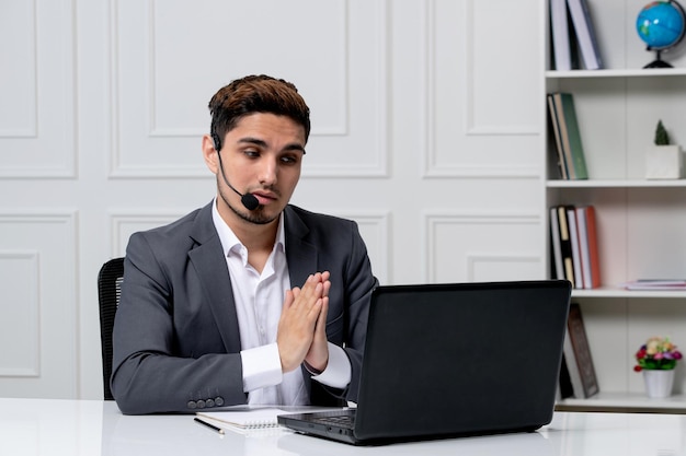 Customer service pretty gentleman with computer in grey office suit holding hands together