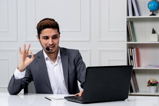 Customer service pretty gentleman with computer in grey office suit showing ok sign