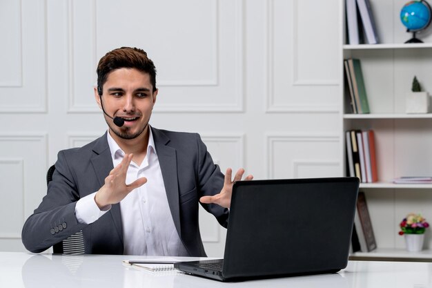 Customer service young cute guy in grey office suit with computer explaining to the client