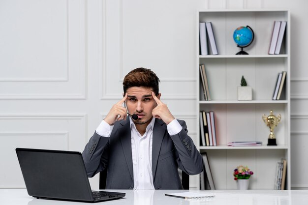 Customer service young cute guy in grey office suit with computer holding temples very irritated