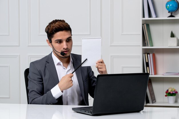 Customer service young cute guy in grey office suit with computer showing notes on the paper