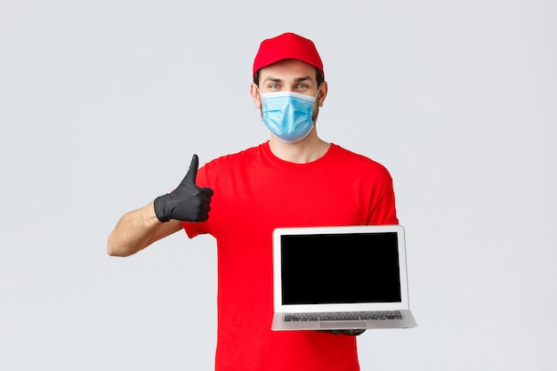 Customer support, covid-19 delivery packages, online orders processing concept. Cheerful courier in red uniform, medical face mask and gloves recommend webpage, show laptop screen and thumb-up
